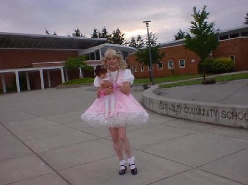 A girl clutching a baby doll while donning a pink satin adult baby dress.